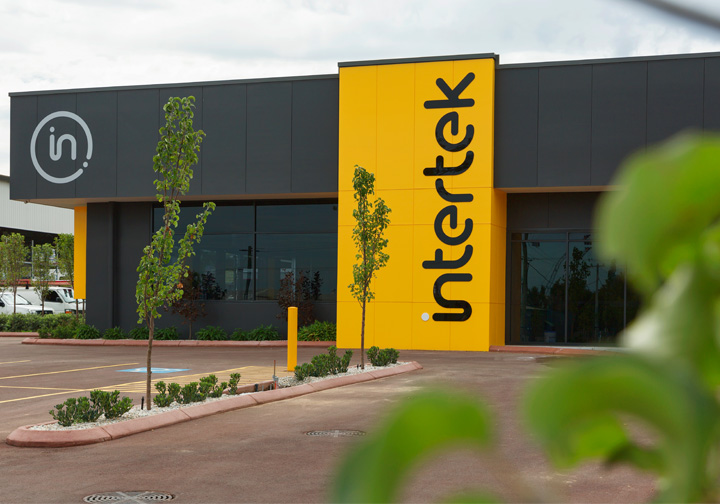 The entrance to the Intertek Minerals Global Centre of Excellence, a dark grey building with a large yellow Intertek sign going vertically down its front. 