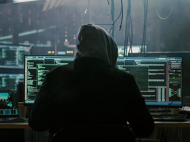 Hooded hacker in dark hideout on a computer with multiple display screens, breaks into data servers with a virus.
