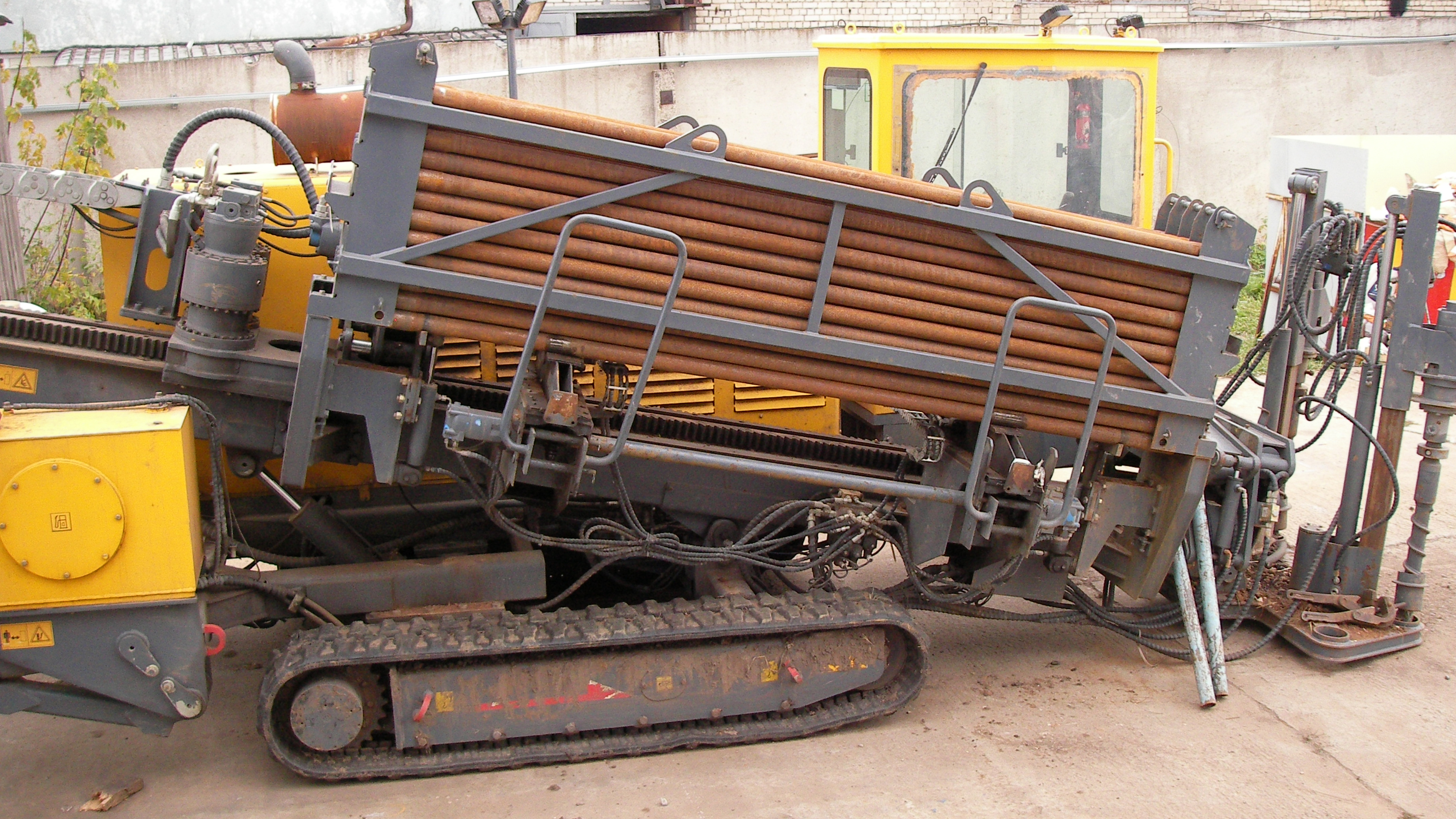 Tracked horizontal directional drilling (HDD) rig parked in an industrial yard, equipped with a rack of steel drill rods mounted on an angled loading system.